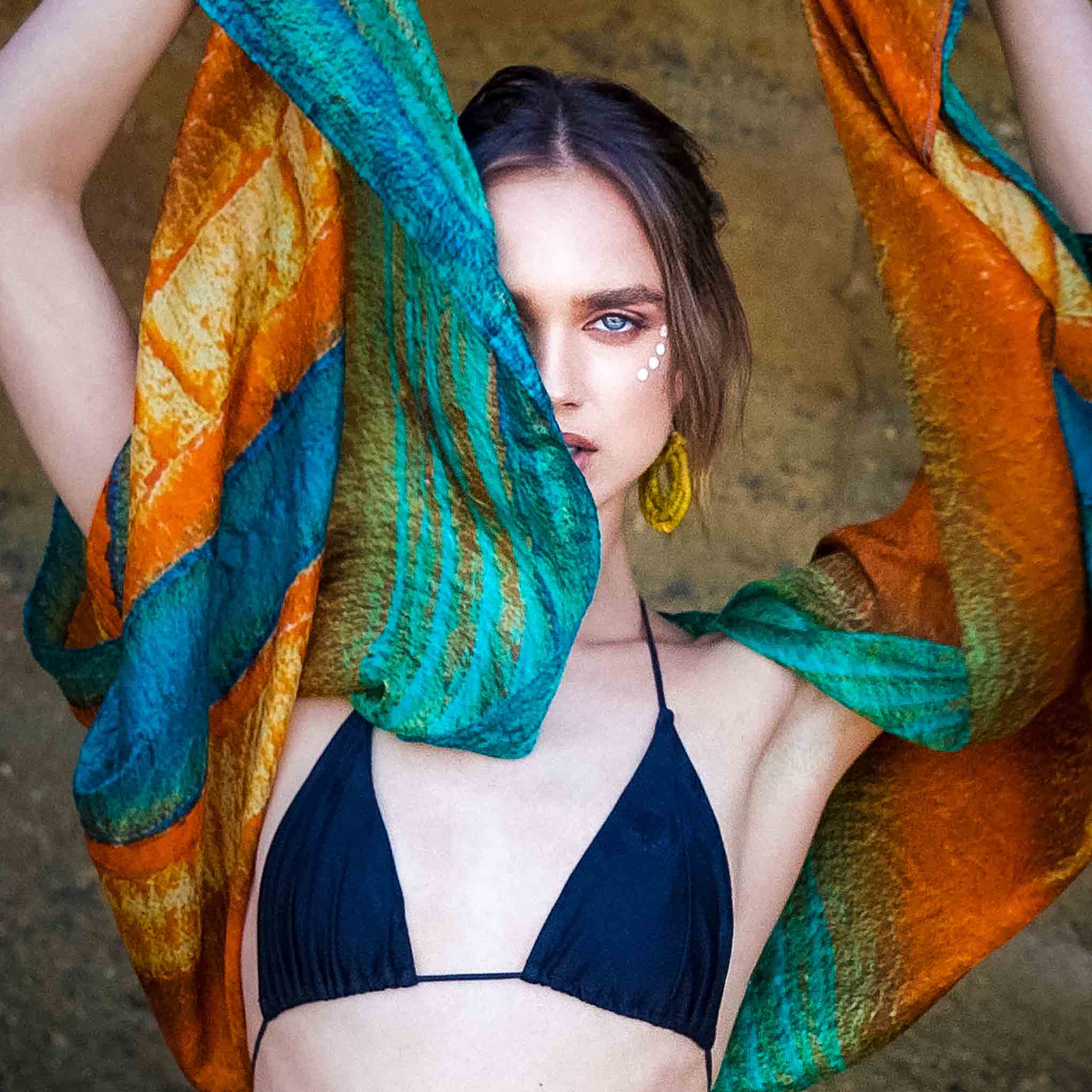Woman in a black bikini with Kamari scarf draped over her shoulders against a textured wall.