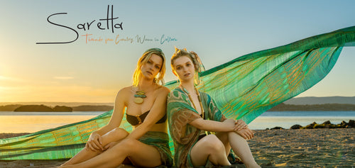 Two women sitting on a beach with a green and gold fabric, sunset in the background, 'Saretta' brand visible.